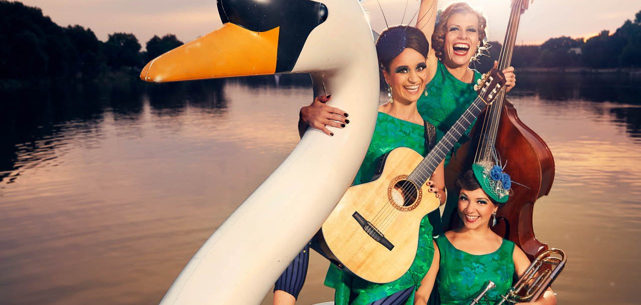 Drei Musikerinnen in leuchtend grünen Retro-Kleidern posieren auf einem großen weißen Schwanen-Tretboot auf einem See. Eine Frau hält eine Gitarre, eine weitere steht mit einem Kontrabass hinter ihr und hebt lachend einen Arm in die Luft, während die dritte mit einer Posaune im vorderen Bereich des Bootes sitzt. Die Frauen tragen auffällige Kopfschmuck-Accessoires und präsentieren sich gut gelaunt im Stil einer nostalgischen Bühnenshow. Im Hintergrund ist die untergehende Sonne über dem ruhigen See zu sehen.