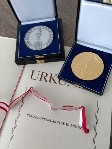 Eine Stadtehrenplakette in Silber und eine Stadtehrenplakette in Gold liegen dekorativ auf einer Urkunde