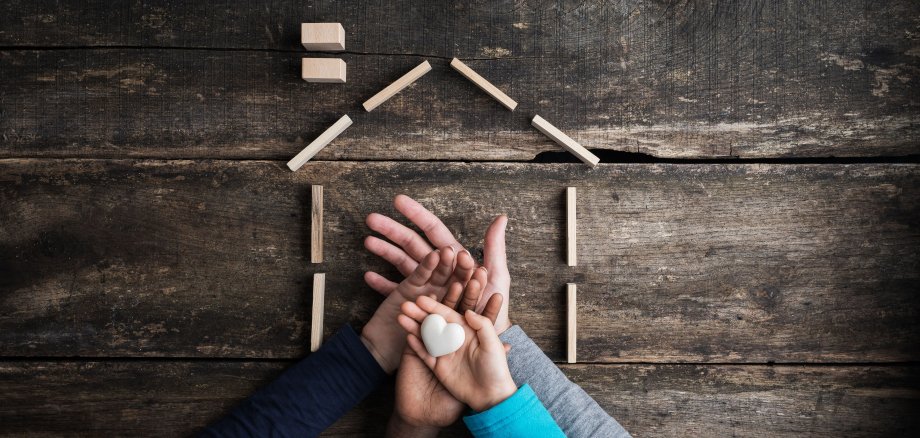 Aufeinander gelegte Hände einer vierköpfigen Familie von oben fotografiert, obere Hand hält Herz und außenherum ein Haus mit Bauklötzen abgebildet 