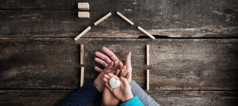 Aufeinander gelegte Hände einer vierköpfigen Familie von oben fotografiert, obere Hand hält Herz und außenherum ein Haus mit Bauklötzen abgebildet 