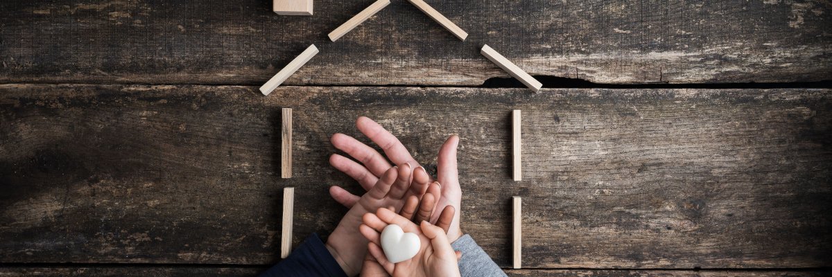 Aufeinander gelegte Hände einer vierköpfigen Familie von oben fotografiert, obere Hand hält Herz und außenherum ein Haus mit Bauklötzen abgebildet 