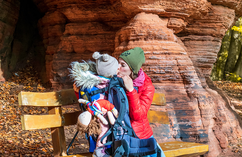 Mama küsst kleines Kind in der Trage vor der Schillerwand